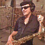 A man in sunglasses playing a saxophone in a recording studio.